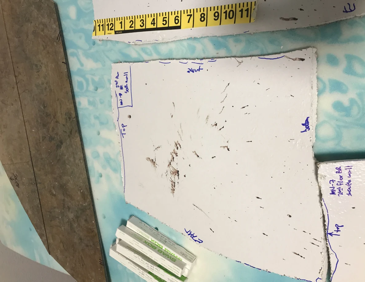 Irregular white wall-panel piece with dark marks, a yellow tape measure and small labeled boxes nearby.