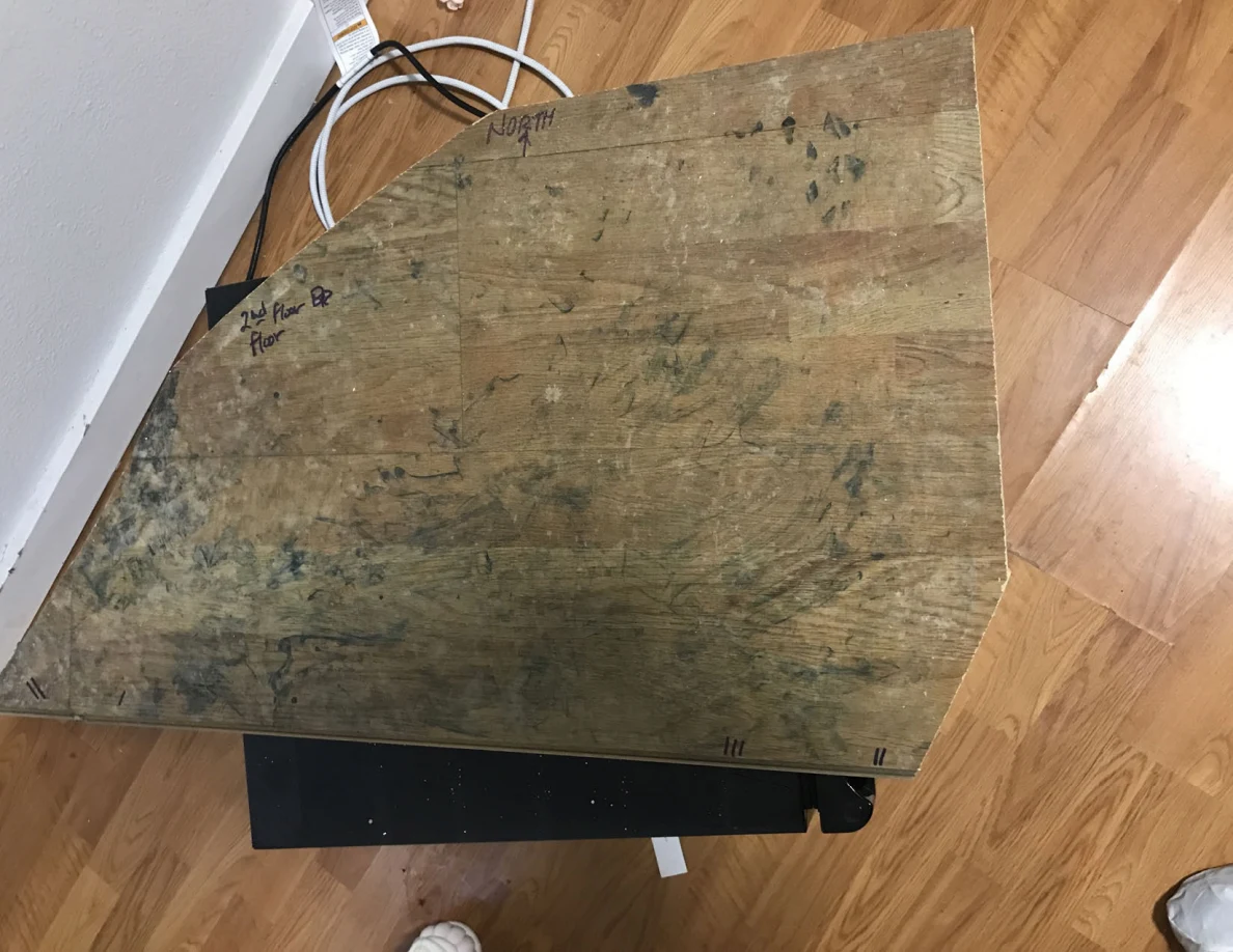 Irregular wooden panel cutout with dark marks and handwritten edge labels on a wood floor.
