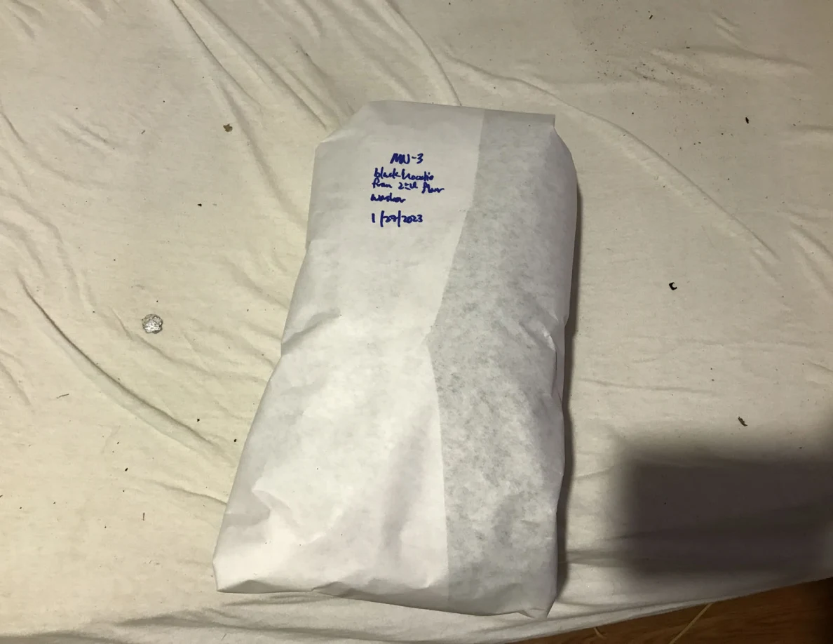 Narrow white-paper-wrapped package with handwritten blue label on a pale sheet.