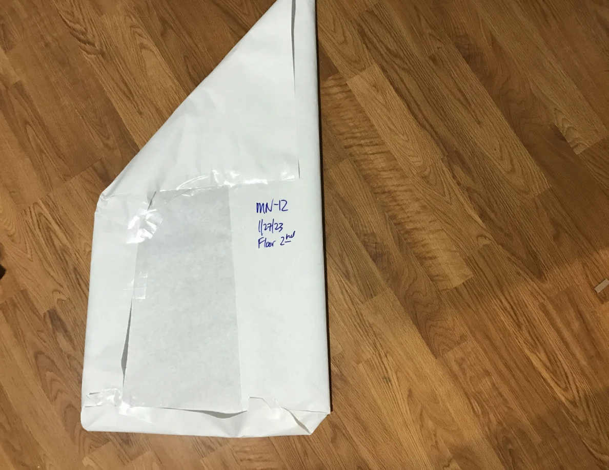 Long angular white-paper-wrapped panel with a handwritten label on a wooden floor.
