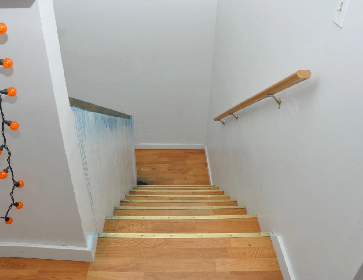 View down wooden stairs with pale walls, wooden handrail and orange lights at left.