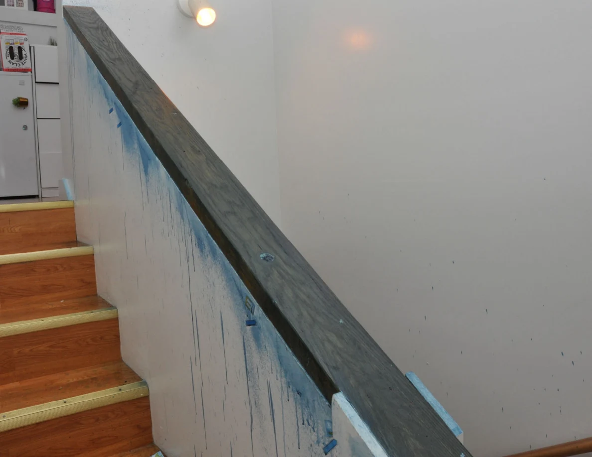 Dark stair cap and blue-streaked half-wall beside wooden stairs and a refrigerator.