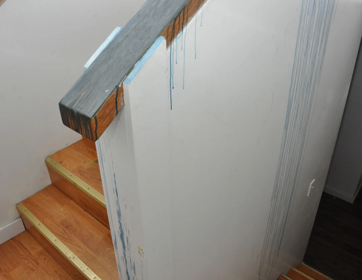Wood-capped half-wall with blue coating and long blue streaks beside wooden stairs.
