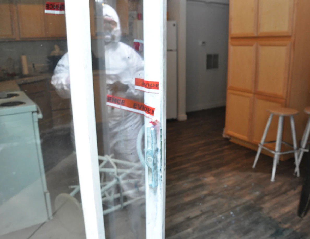 Sliding glass doorway with red evidence tape, kitchen stools and reflection of a person in protective coveralls.