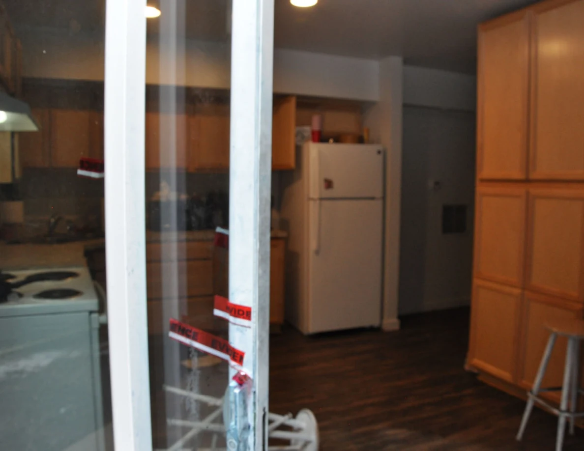 View through a sliding glass door into a kitchen with refrigerator, wooden cabinets and stools.