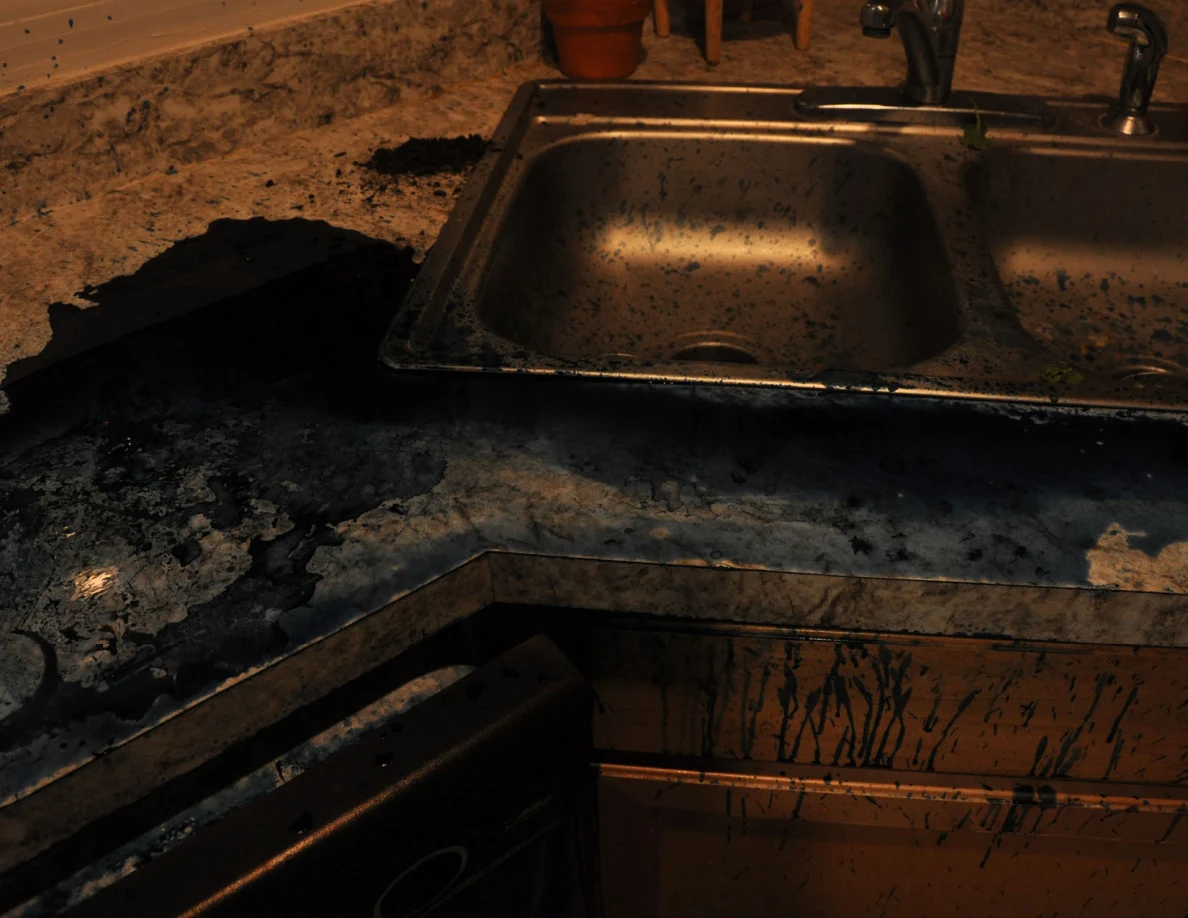 Dim view of a kitchen sink and countertop with dark blue residue and streaked cabinet fronts.
