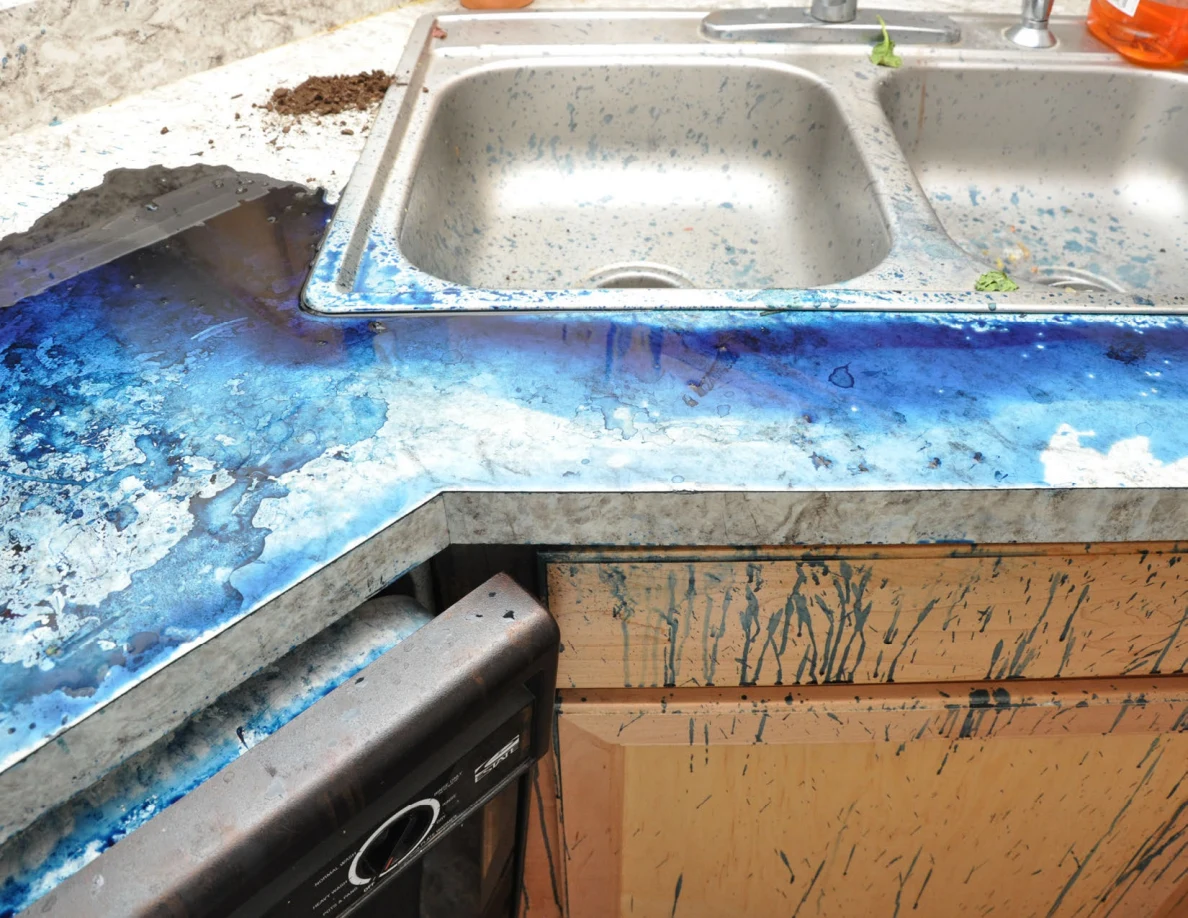 Stainless-steel sink beside blue-covered countertop and streaked wooden cabinets.