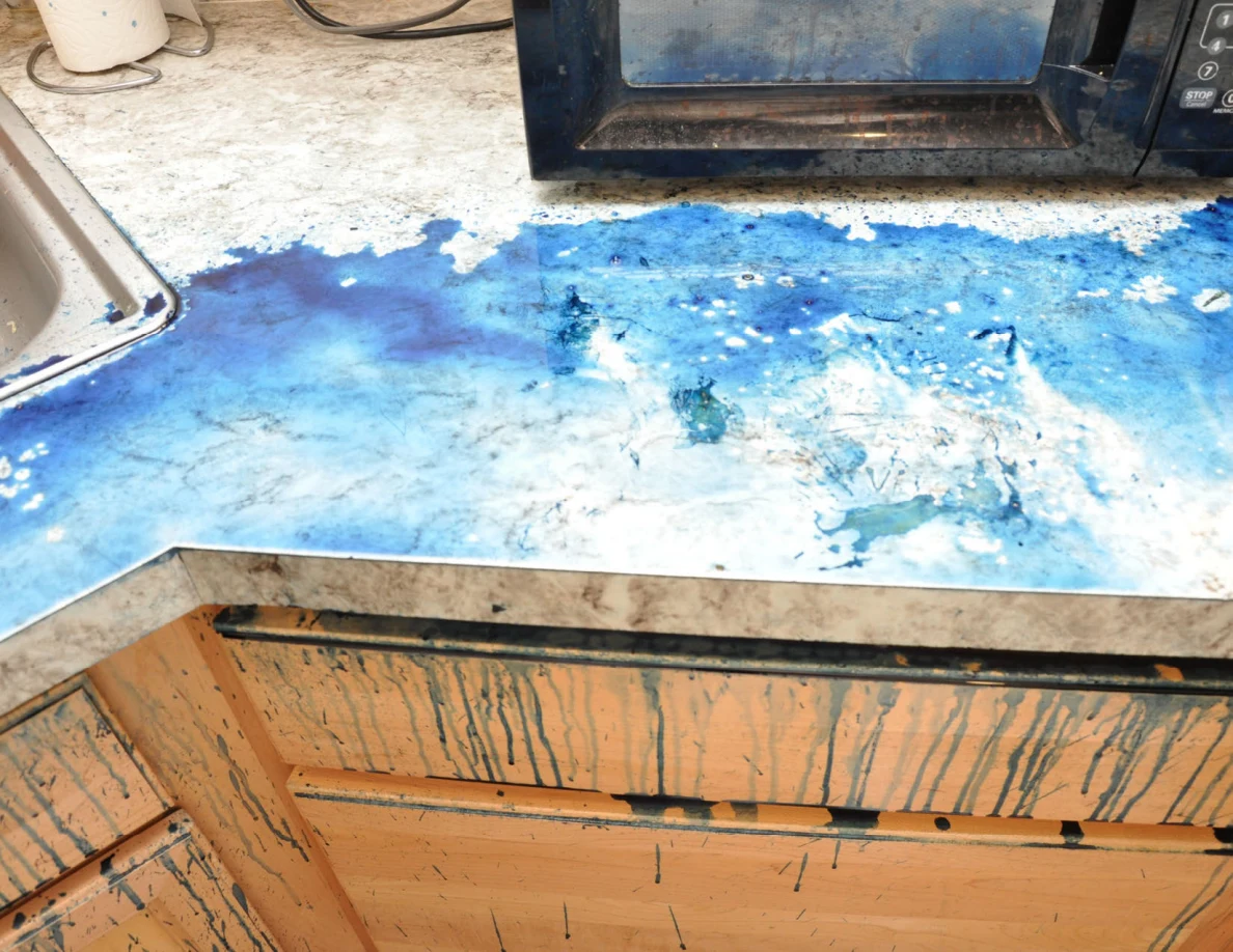 Blue-covered counter beneath a microwave, above streaked wooden drawers.