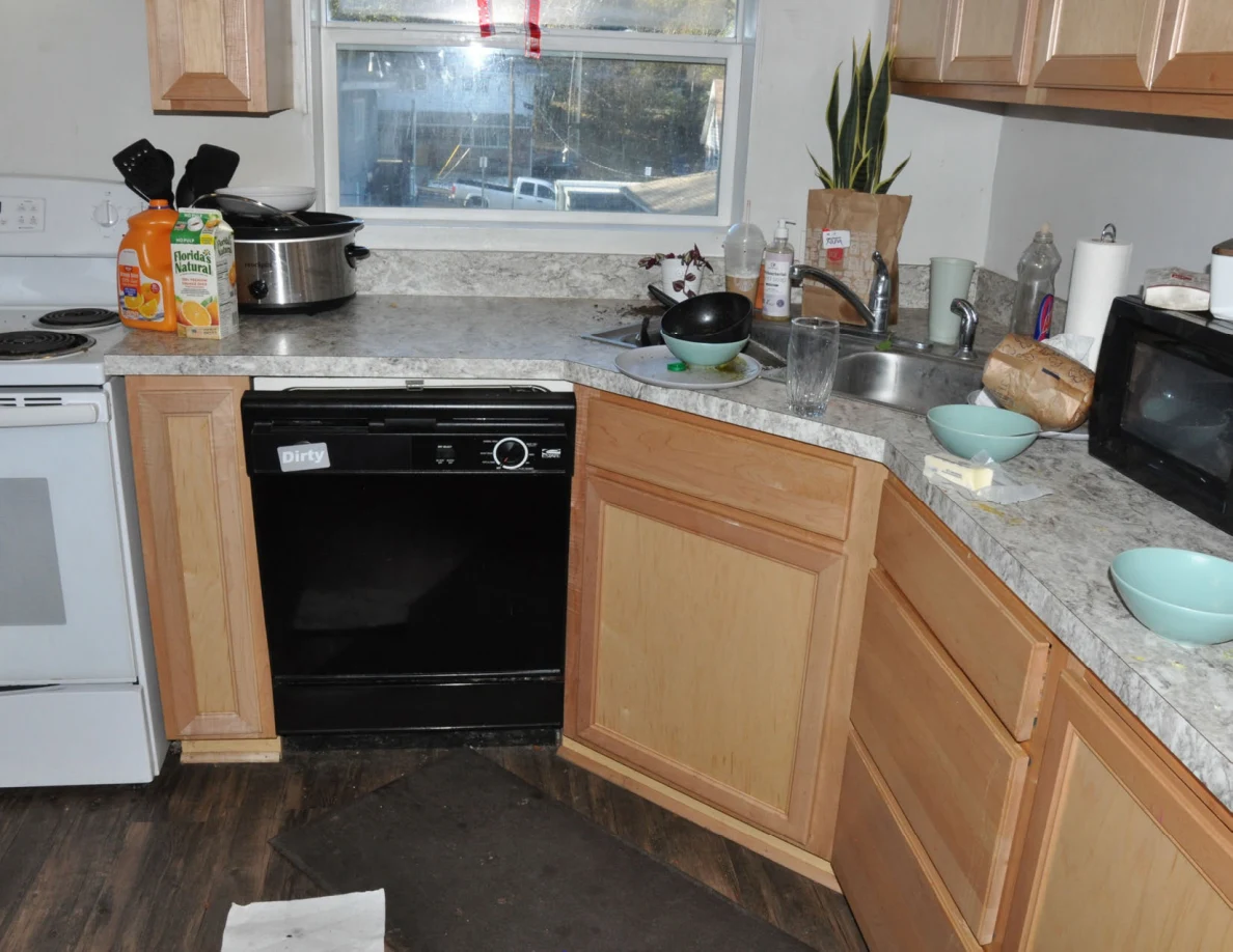 Kitchen sink, dishwasher and wooden cabinets with dishes, slow cooker and drink containers on the counter.
