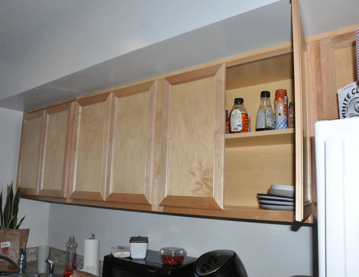 Upper wooden kitchen cabinets with one door open, showing bottles and stacked plates.