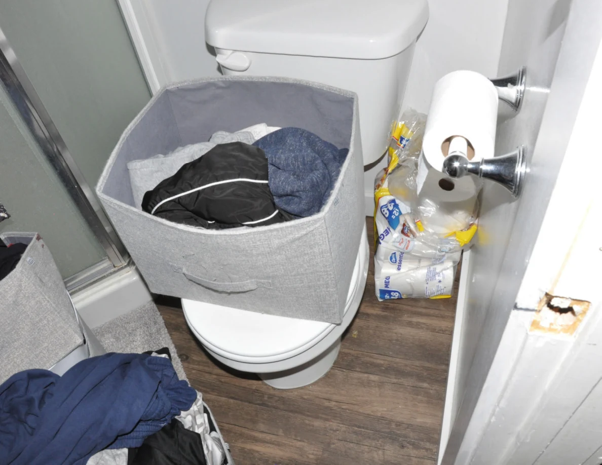 Gray fabric bin containing clothing on a toilet seat beside toilet-paper rolls.