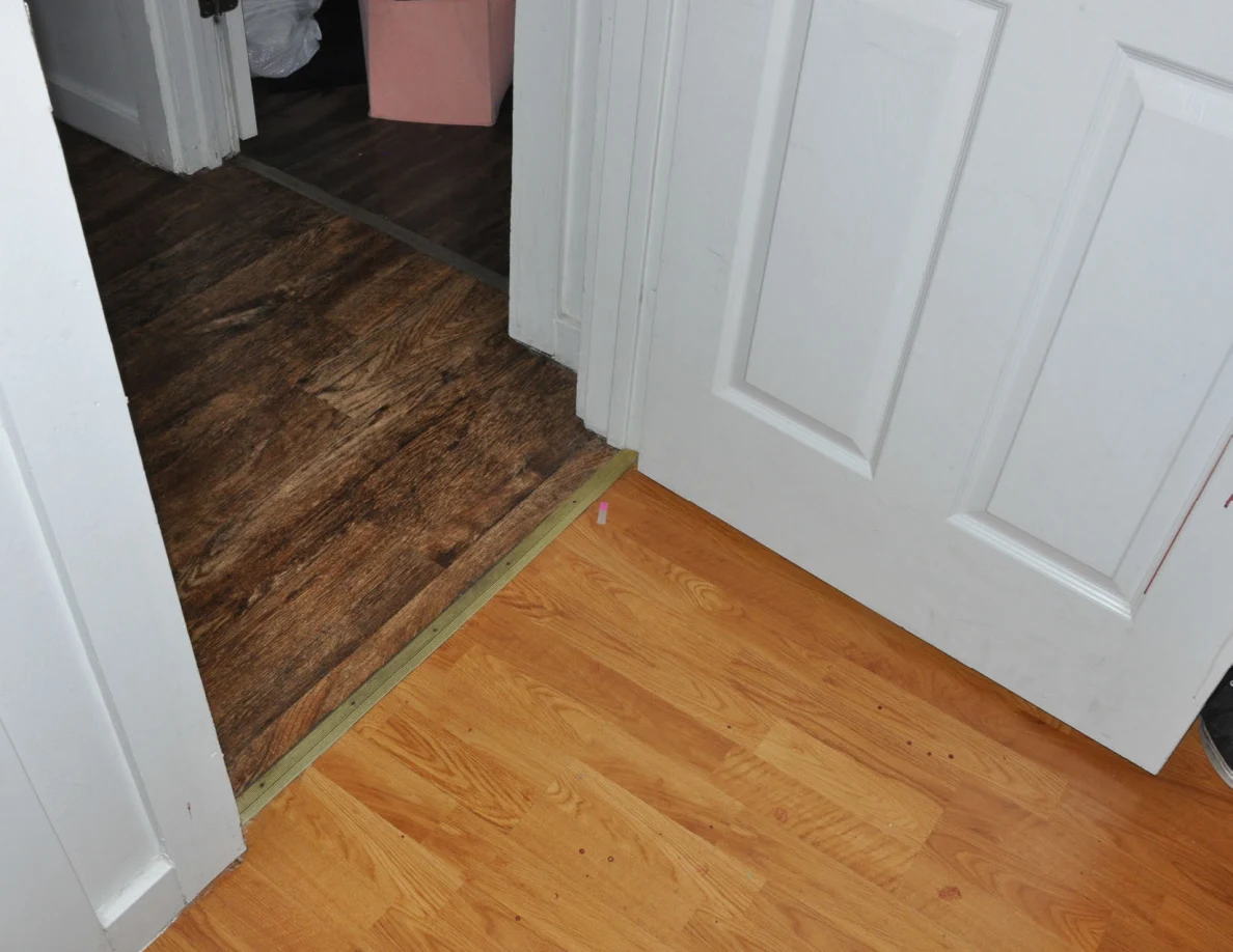 Open white doorway between two wood-look floor finishes, with clothing bins in the next room.