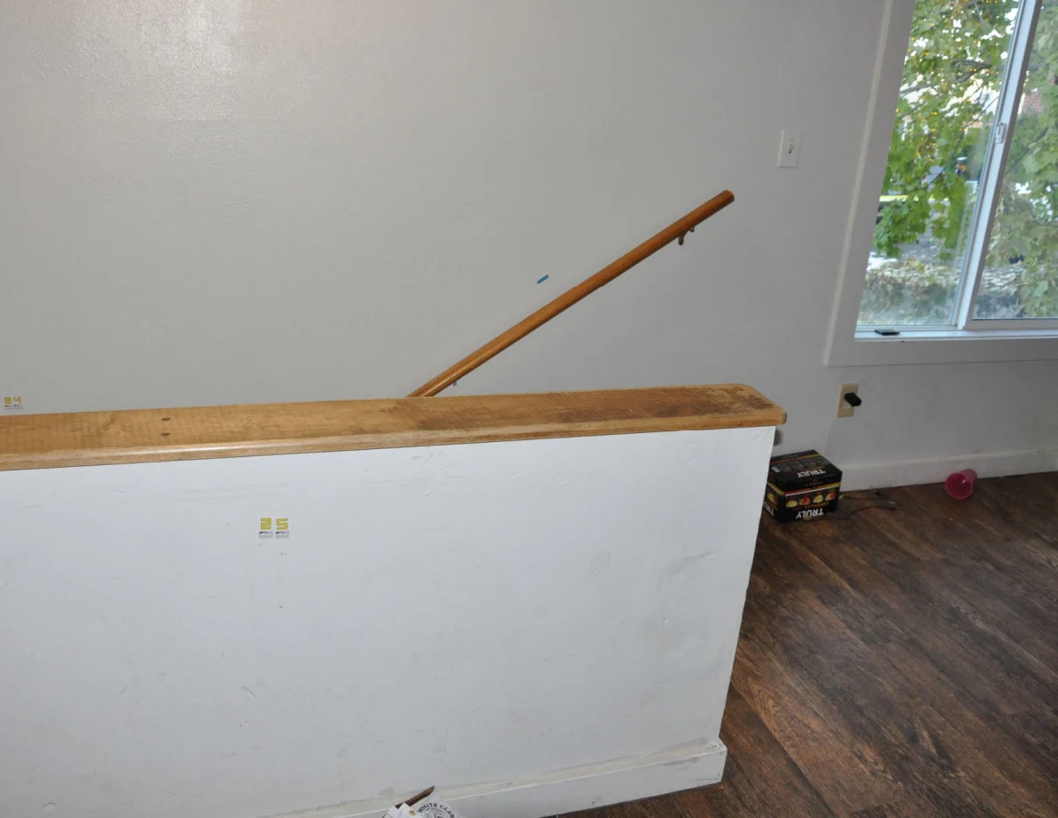 Stair half-wall beneath a wooden handrail, with a window and drink packaging nearby.
