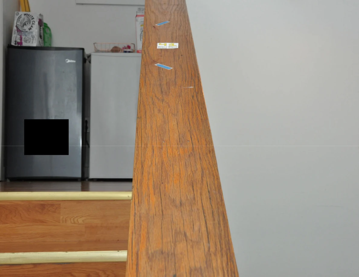 Wooden stair cap with yellow label and blue arrow stickers, with small refrigerators beyond and a black redaction.