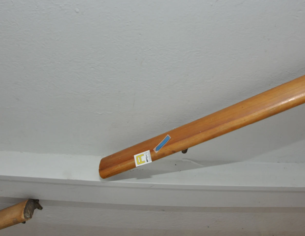 Lower end of a wooden handrail with a letter label and blue arrow sticker.