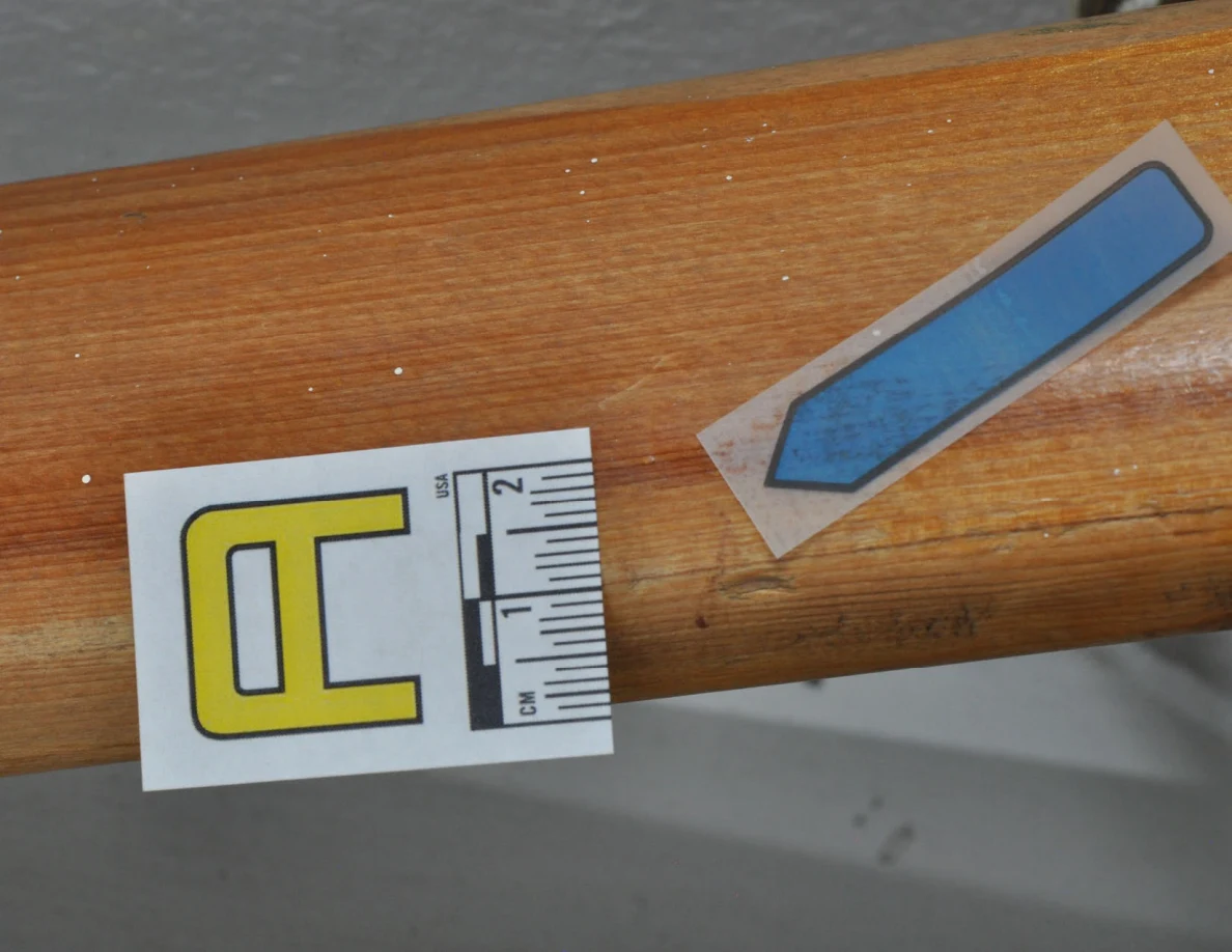 Close view of a letter label and blue arrow beside a dark mark on wood grain.