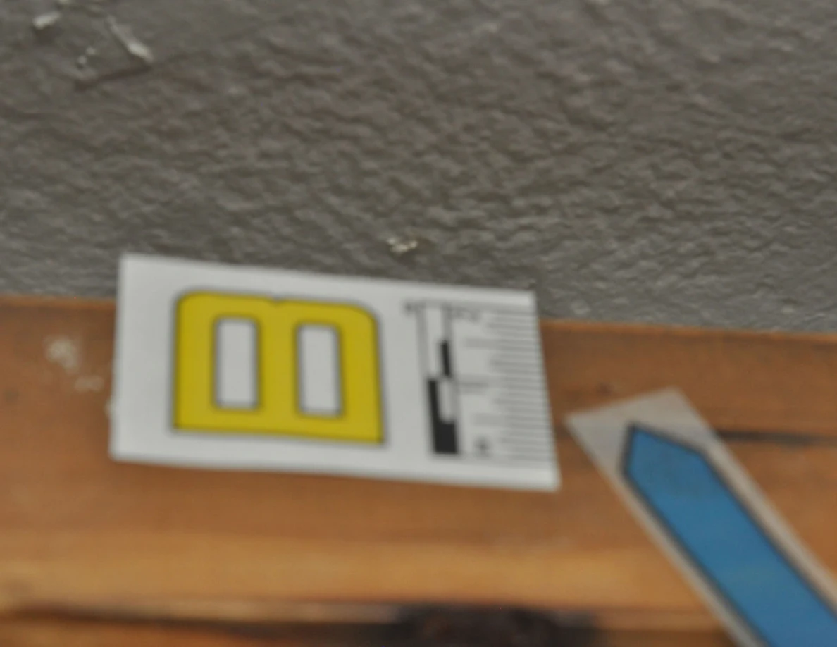 Close view of label B and blue arrow on a wooden handrail beneath scuffed wall paint.