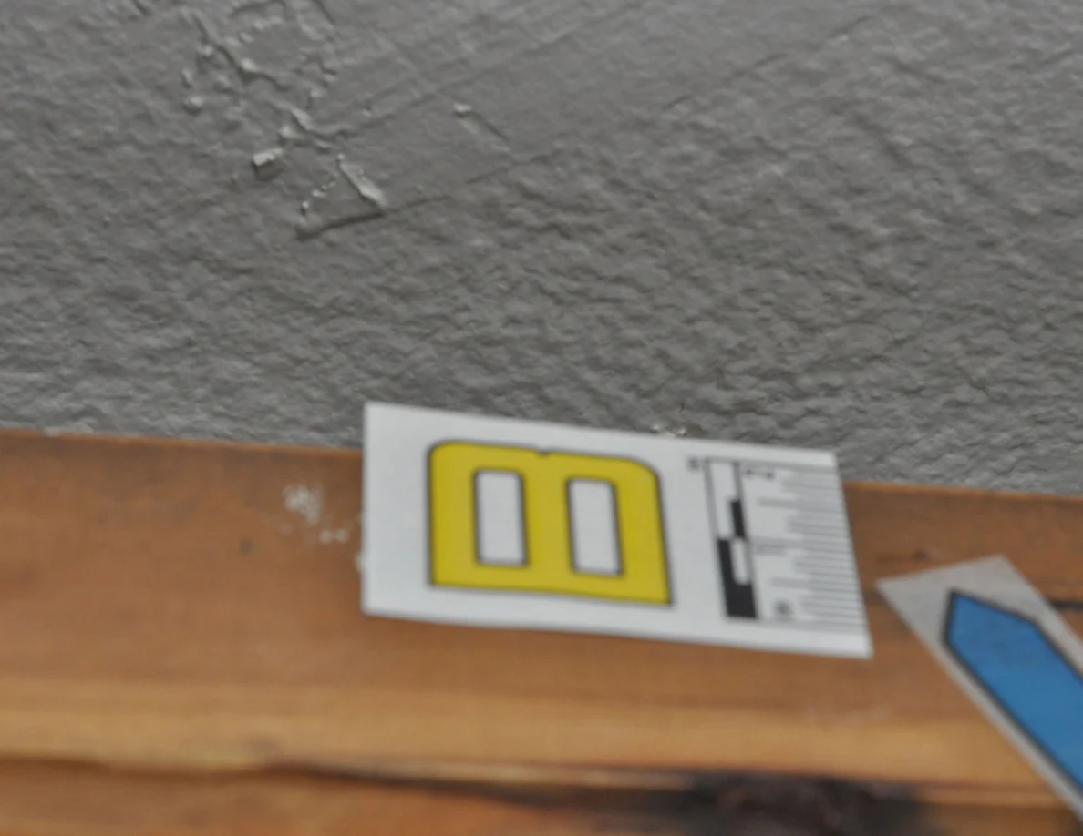 Letter label B and blue arrow on wood beneath a patch of cracked wall paint.
