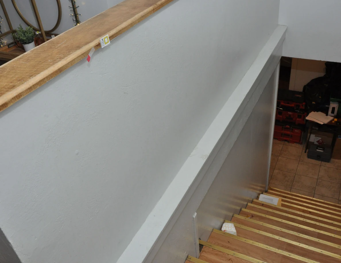 View down stairs beside a wood-capped half-wall with a small letter label and pink tape.