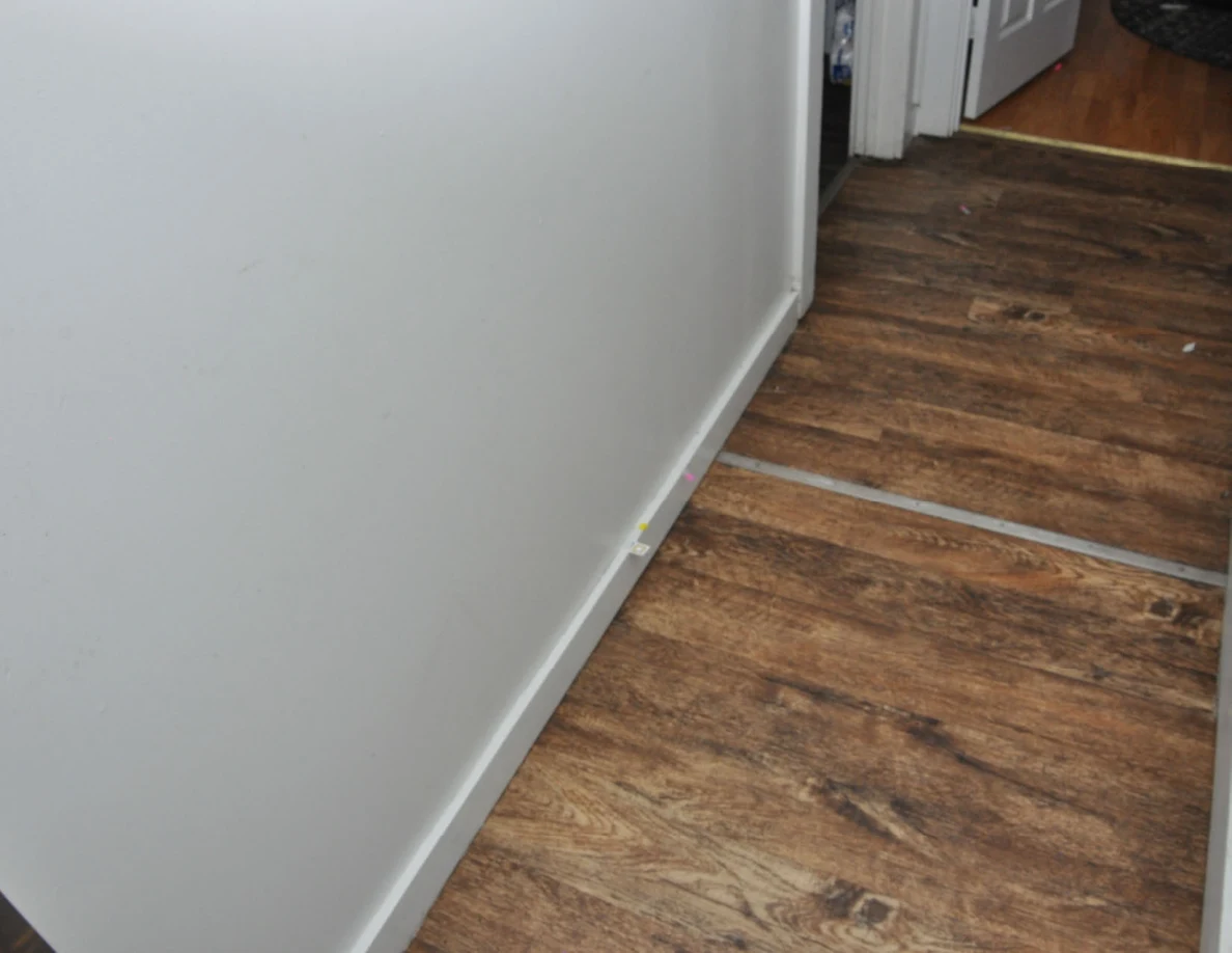 Hallway wall and white baseboard with small tape markers beside wood-look flooring.