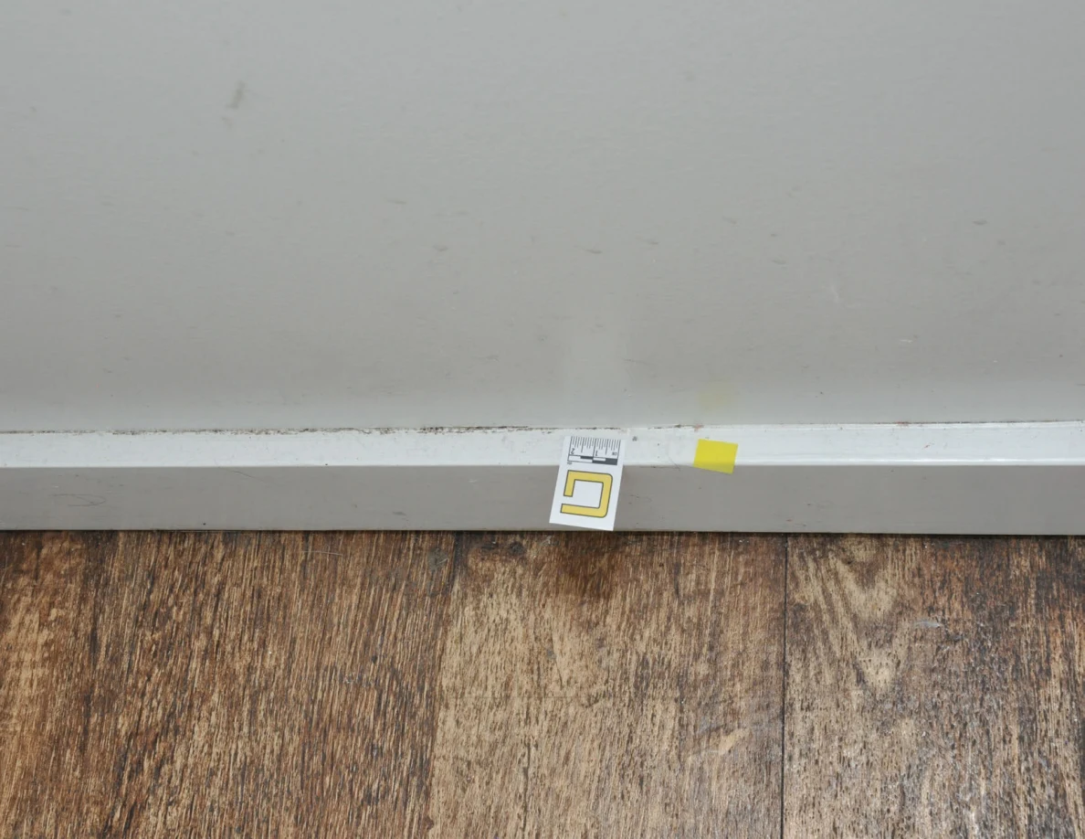 Close view of a baseboard letter label and yellow tape below faint wall marks.