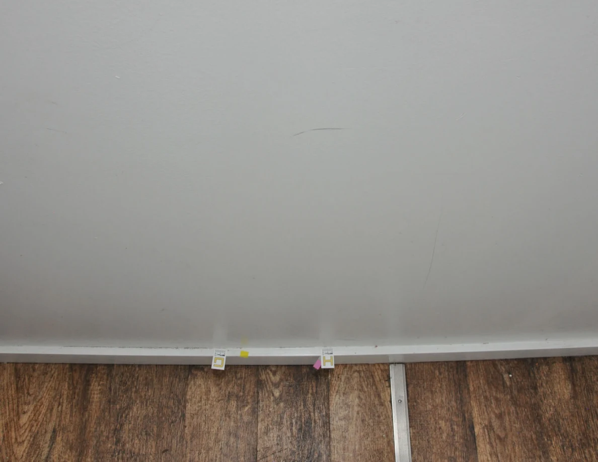 Lower wall and baseboard with two letter labels and colored tape beside a metal floor strip.