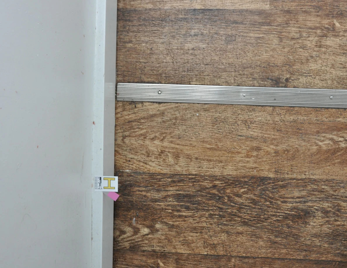 Rotated view of a baseboard letter label and pink tape beside wood-look flooring.