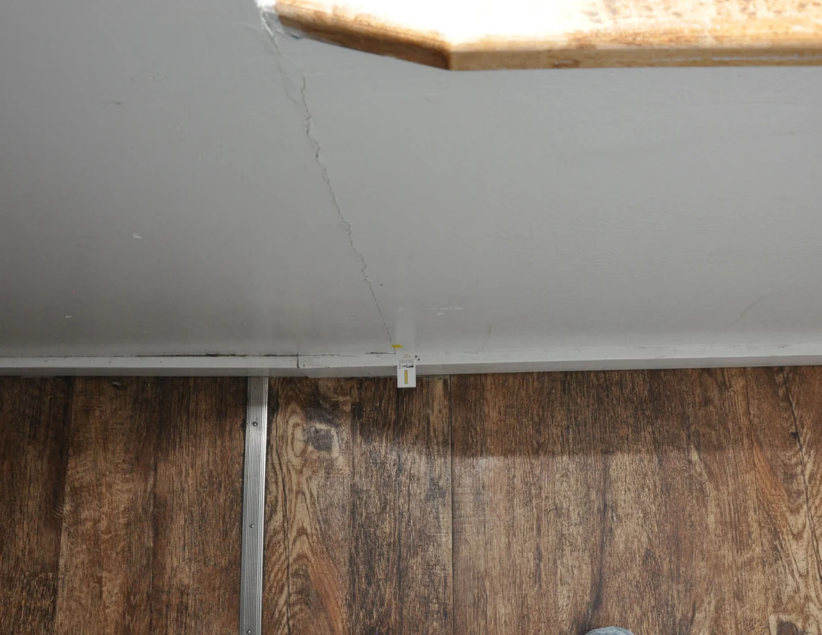 Vertical crack running down a half-wall beneath the end of its wooden cap toward a baseboard label.