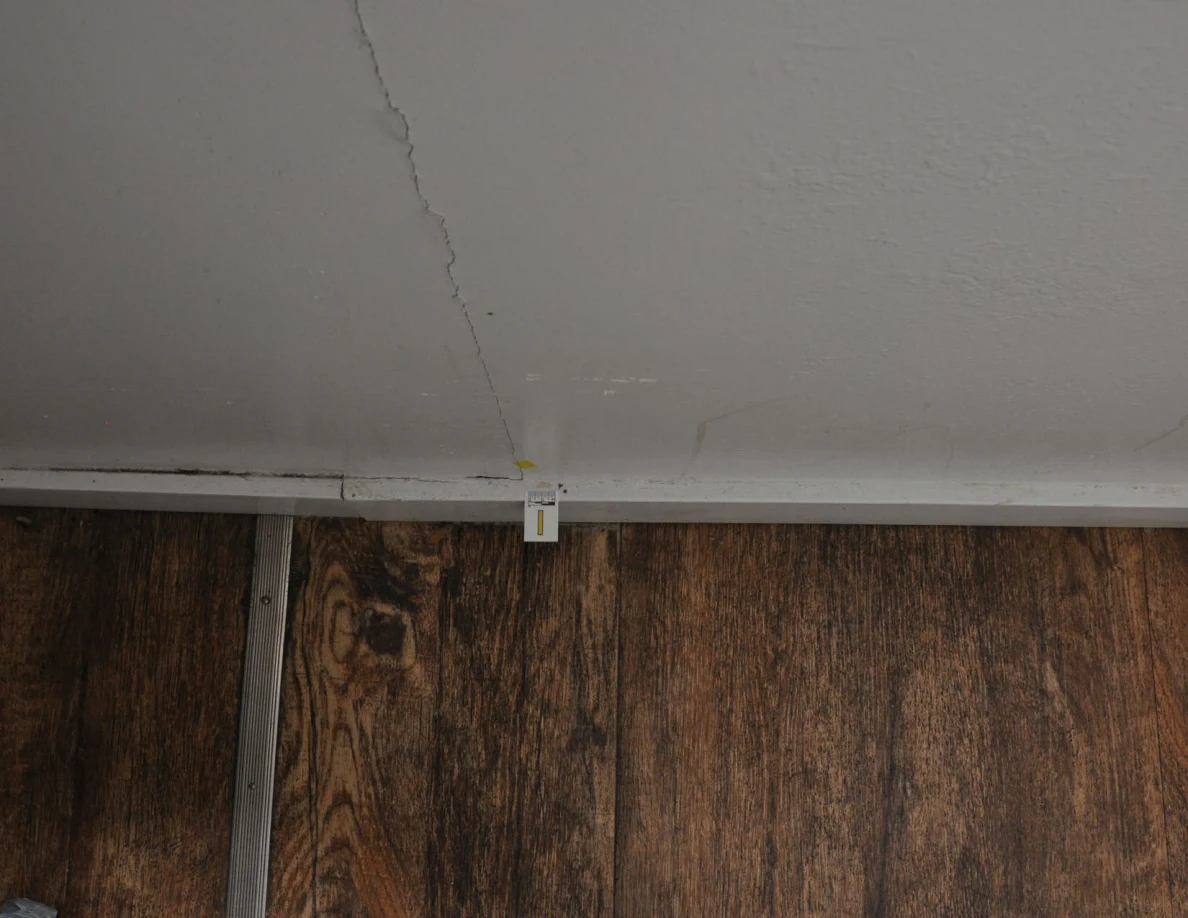 Close view of a vertical wall crack above a small baseboard label and yellow tape.