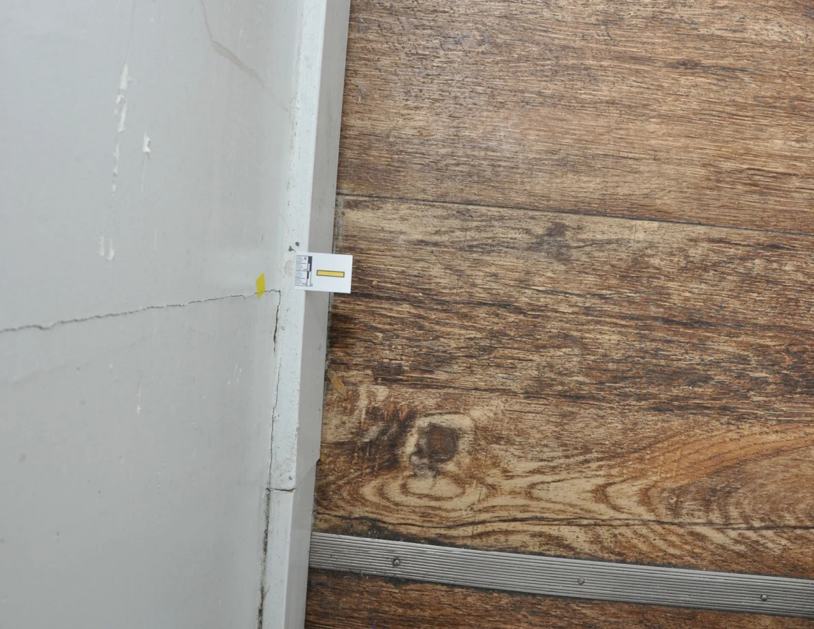 Rotated view of a cracked wall and baseboard with a small letter label beside wood-look floor.