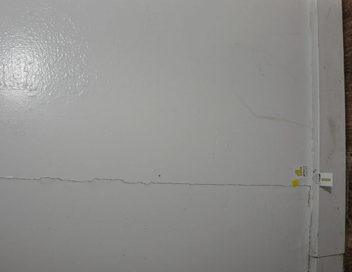Rotated close view of a wall crack beside small labels and yellow tape near a baseboard.