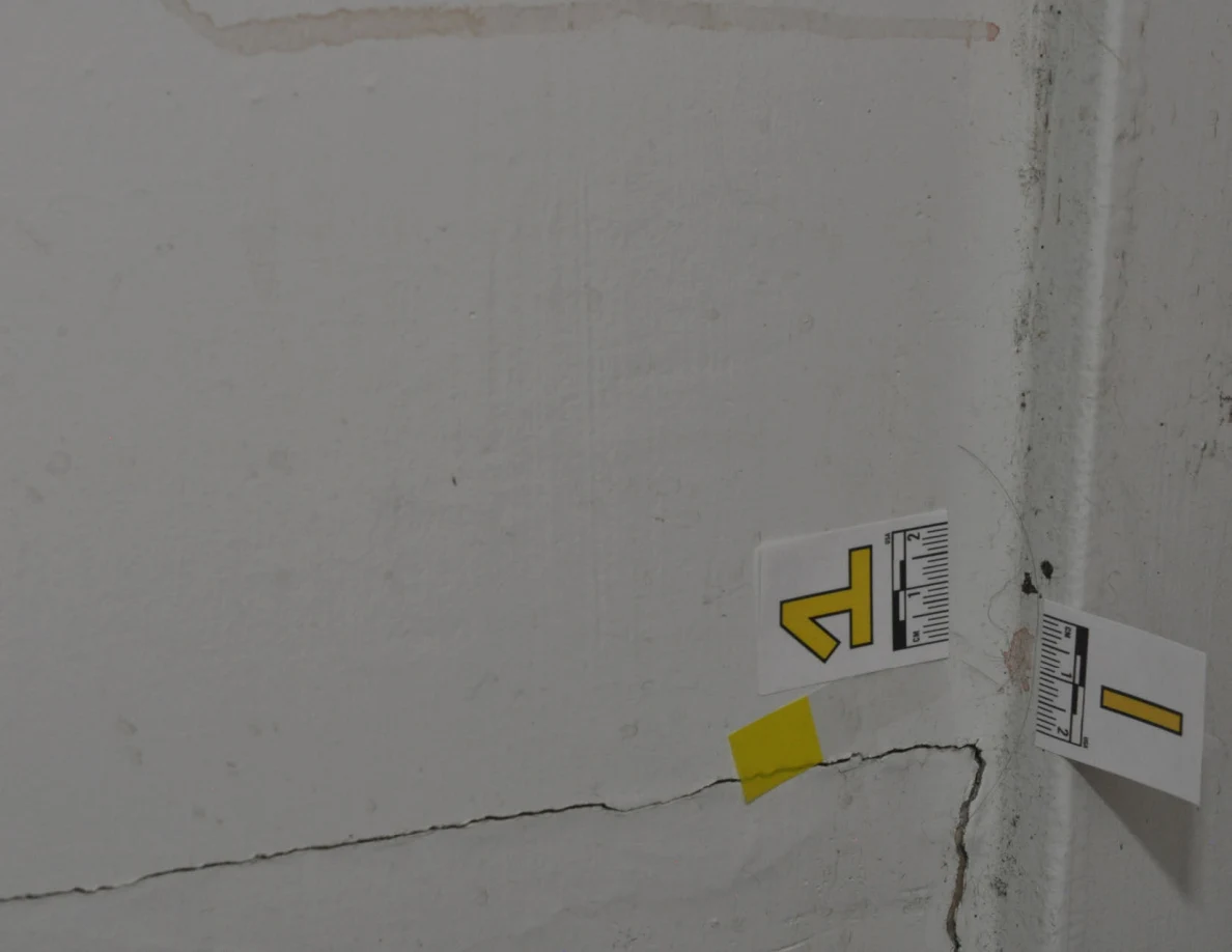 Rotated view of a wall crack, scuffed baseboard and two letter labels with yellow tape.