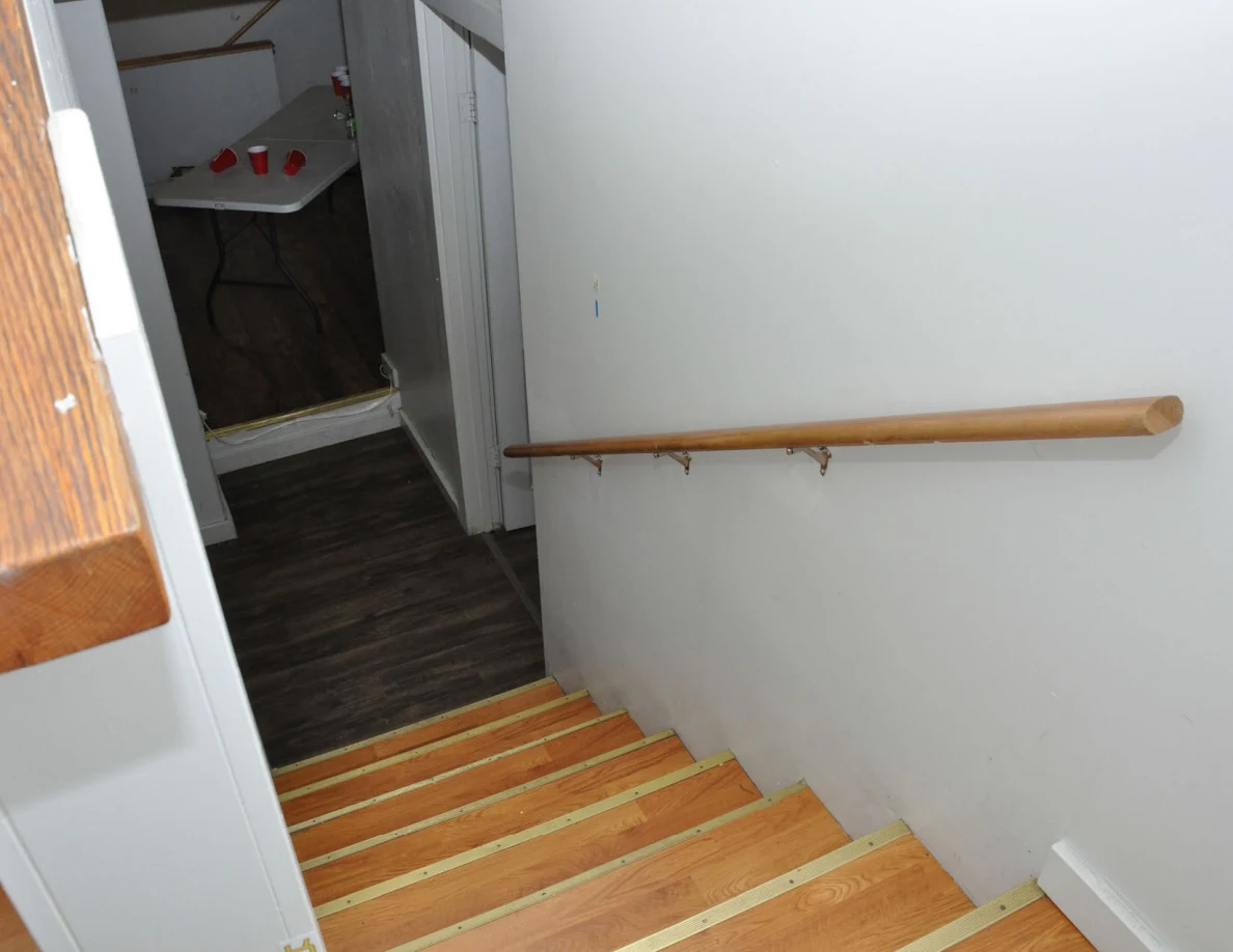 View down wooden stairs beside handrail toward a hallway and table with red cups.