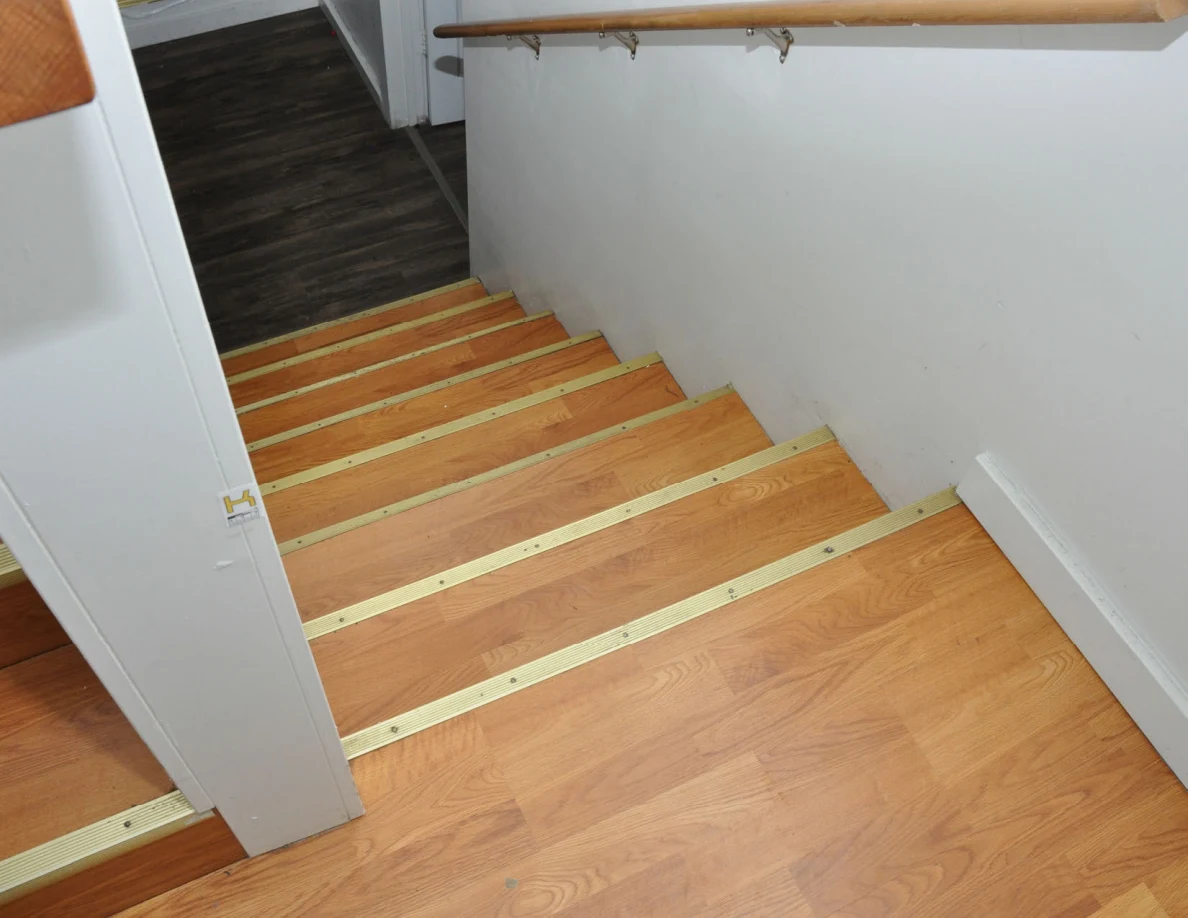 Wooden stairs with metal edges beside pale wall and a small letter label on the half-wall end.