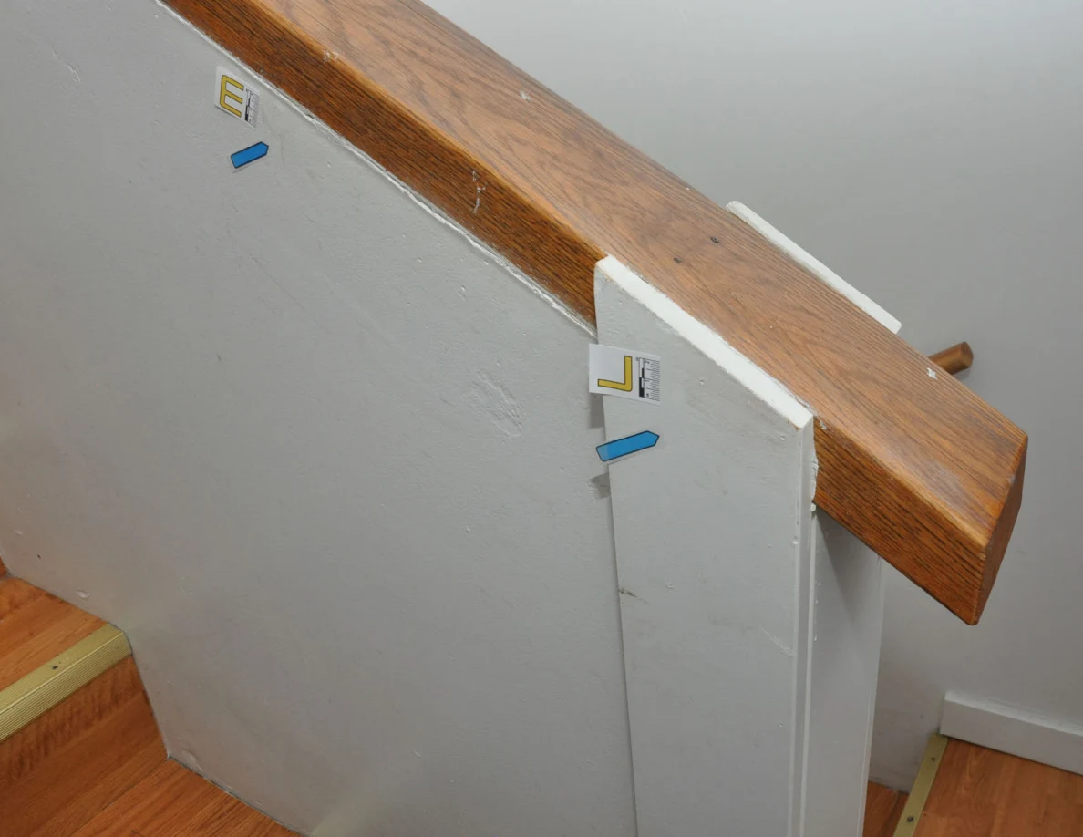 End of wooden stair cap above a pale half-wall with labels and blue arrows.