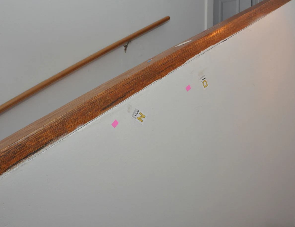 Angled view of stair half-wall with two letter labels and pink tape beneath wooden cap.