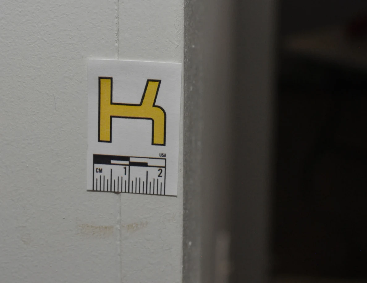 Close view of label K above a faint tan mark on a pale wall edge.