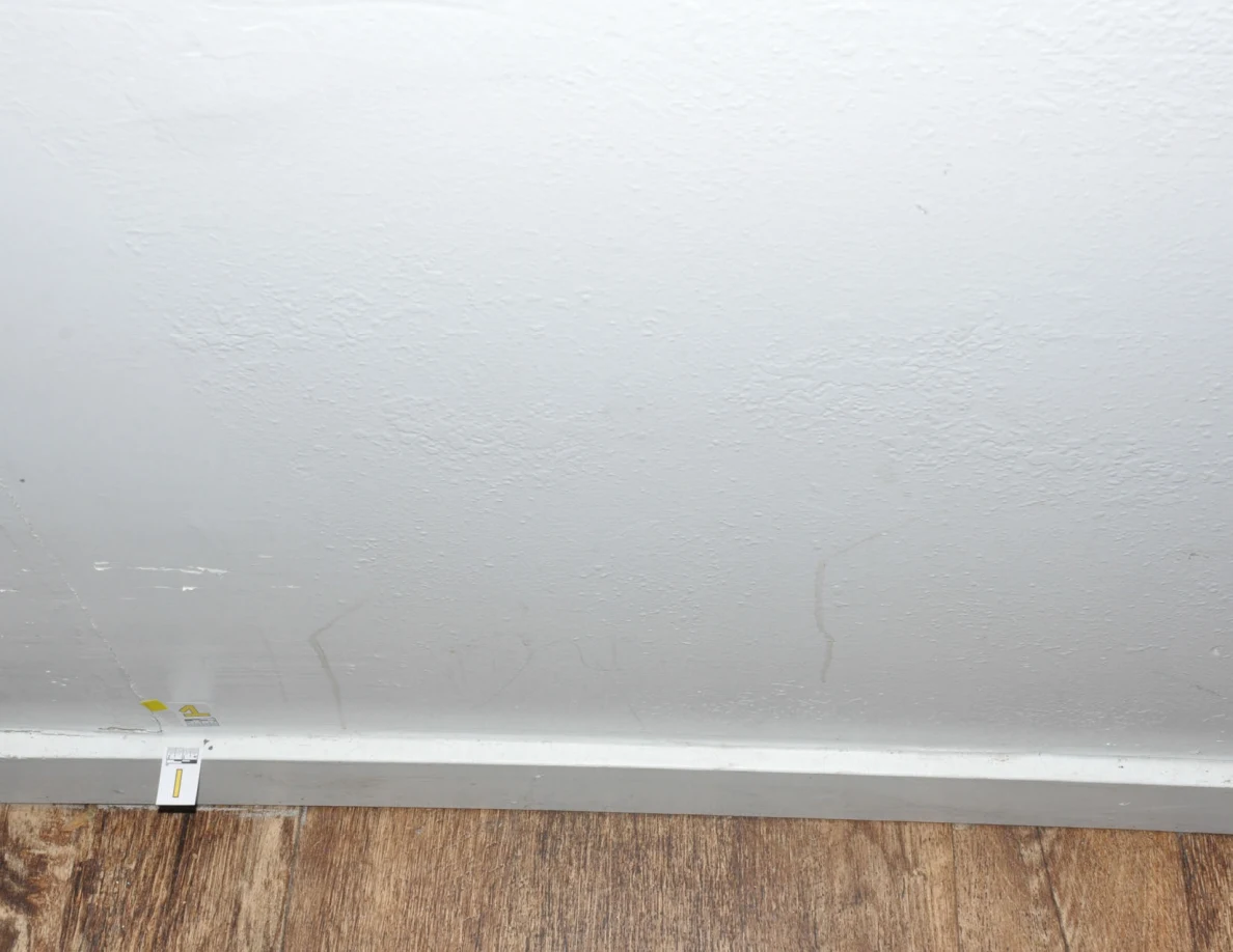 Lower pale wall with faint streaks and a baseboard label beside wood-look floor.