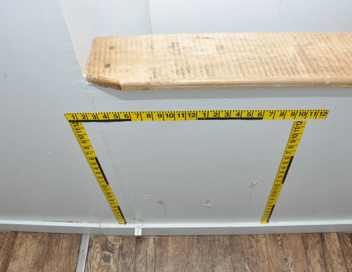 Yellow measurement tape outlining a wall area beneath a wooden cap, with small letter labels.