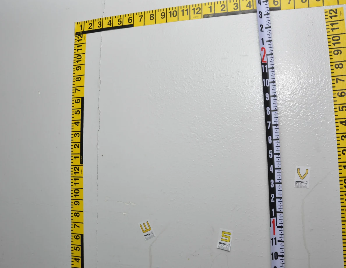 Yellow measurement tape around a wall area with a vertical black-and-white measuring rod near label V.