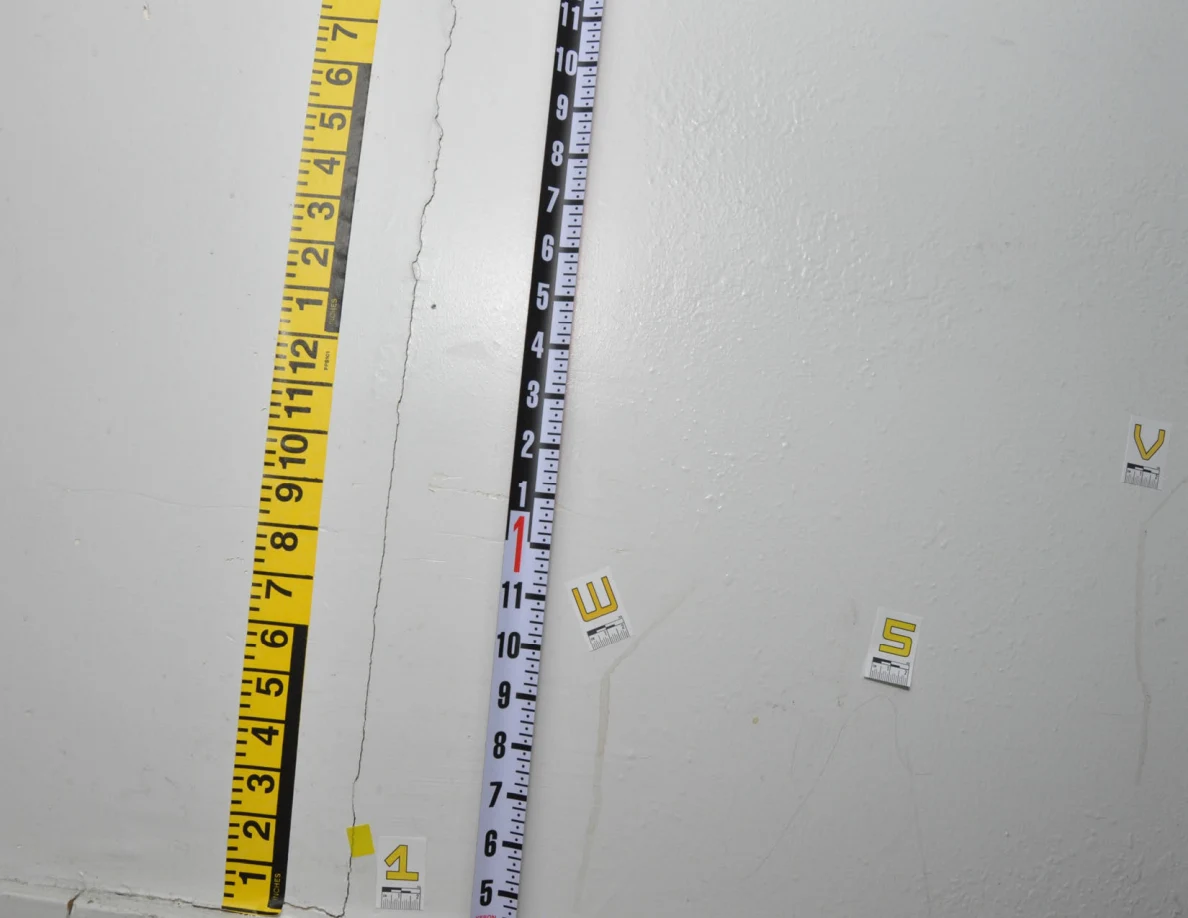 Angled view of a vertical black-and-white measuring rod beside yellow measurement tape and wall labels.