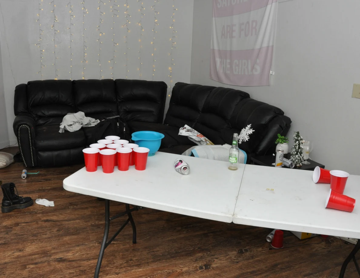 Living room with black sofas, string lights and a white cup-pong table holding red plastic cups, a blue bowl, bottle and crushed can.