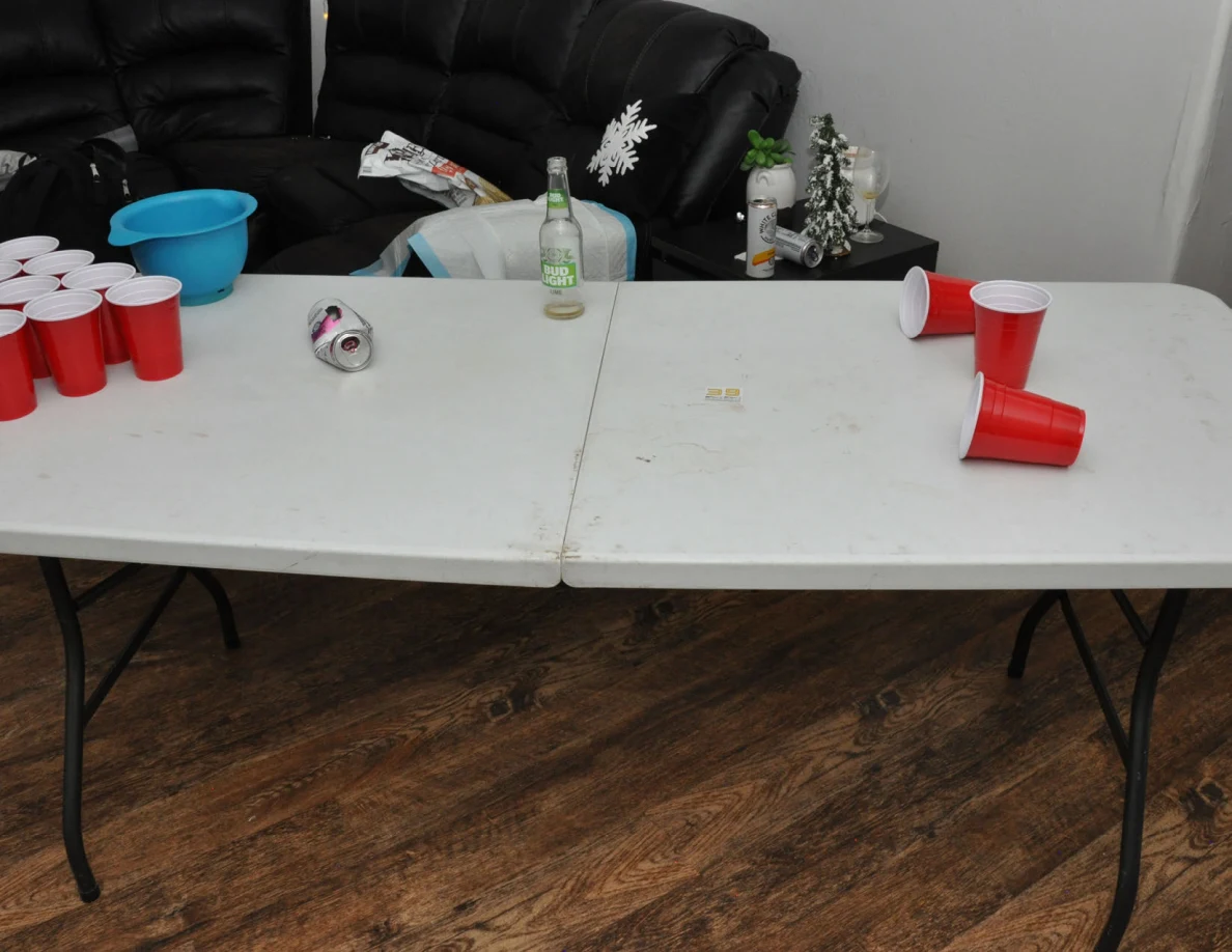 White folding cup-pong table with red plastic cups, a blue bowl, bottle and crushed can in front of black sofas.