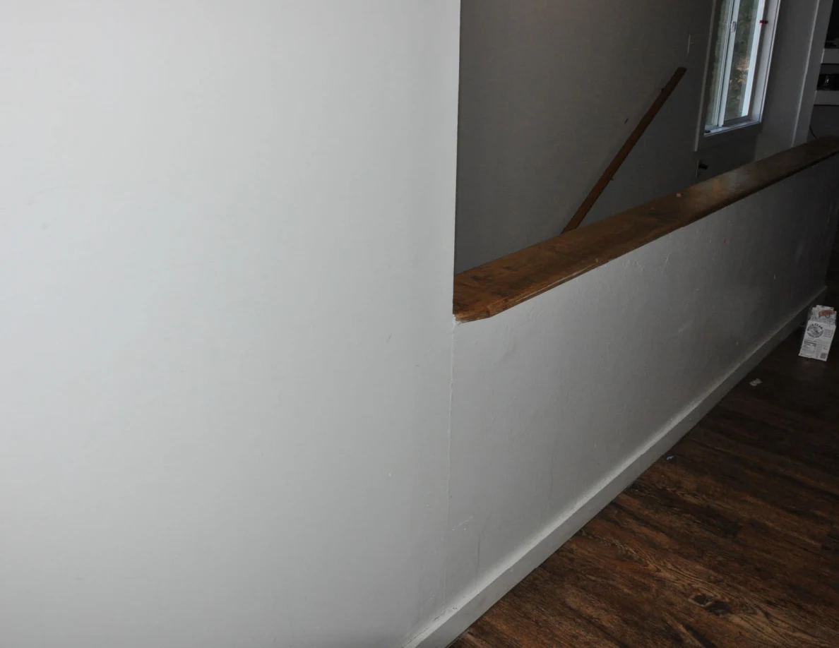 White wall beside a stairwell opening with a wooden rail and wood-pattern floor.