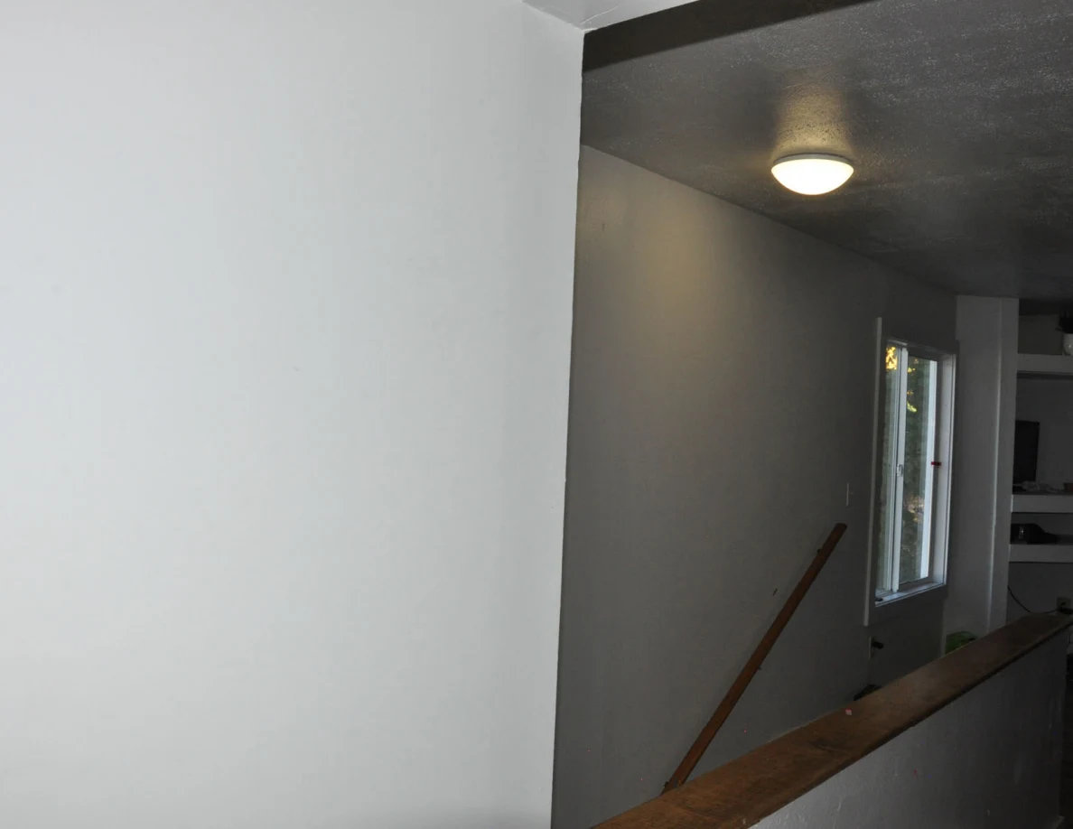 Stairwell opening with white walls, wooden handrail, window and lit ceiling fixture.