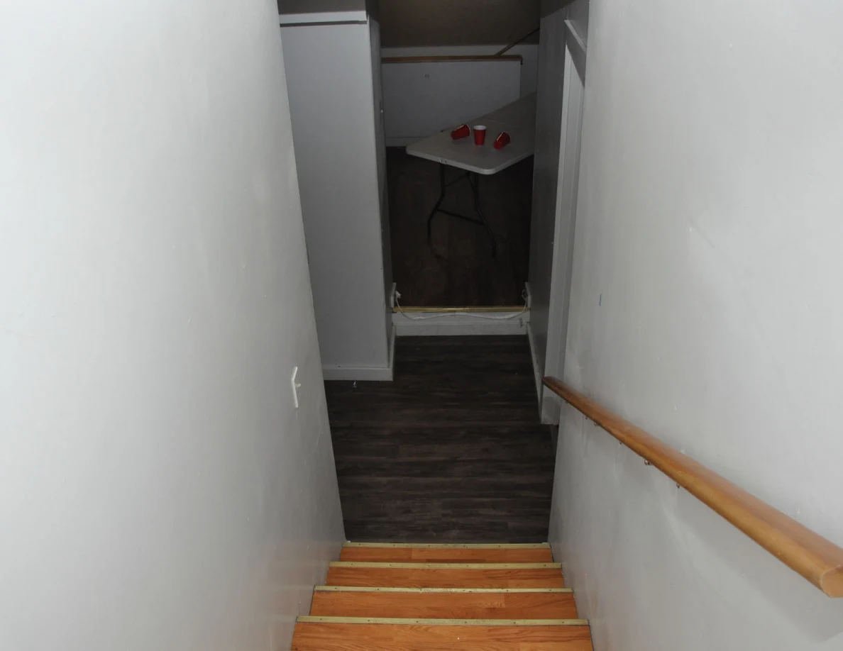 View down wooden stairs with a handrail toward a room containing a white table and red cups.