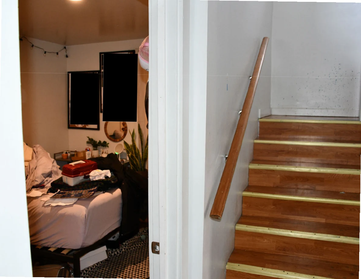 Bedroom doorway beside wooden stairs and handrail, with bedding and objects visible inside.