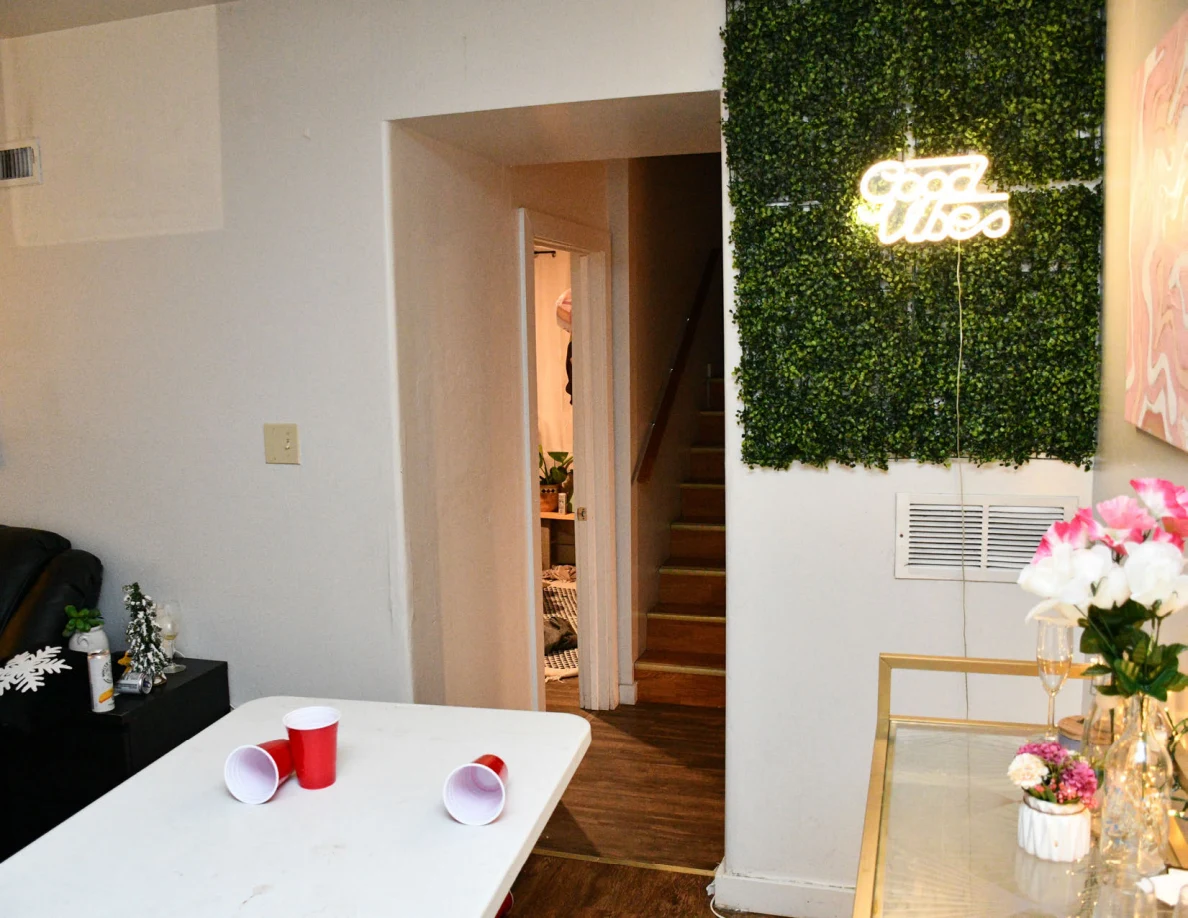 Living area with red cups on a white table, flower vases and a glowing sign mounted on an artificial greenery panel beside stairs.