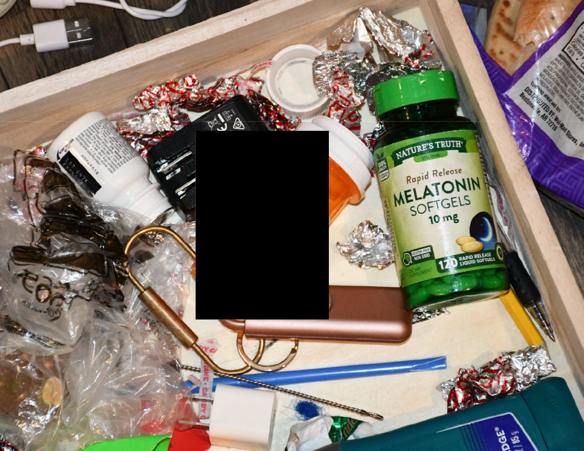 Drawer containing a melatonin bottle, wrappers, eyeglasses, cords and small objects, with a black redaction.