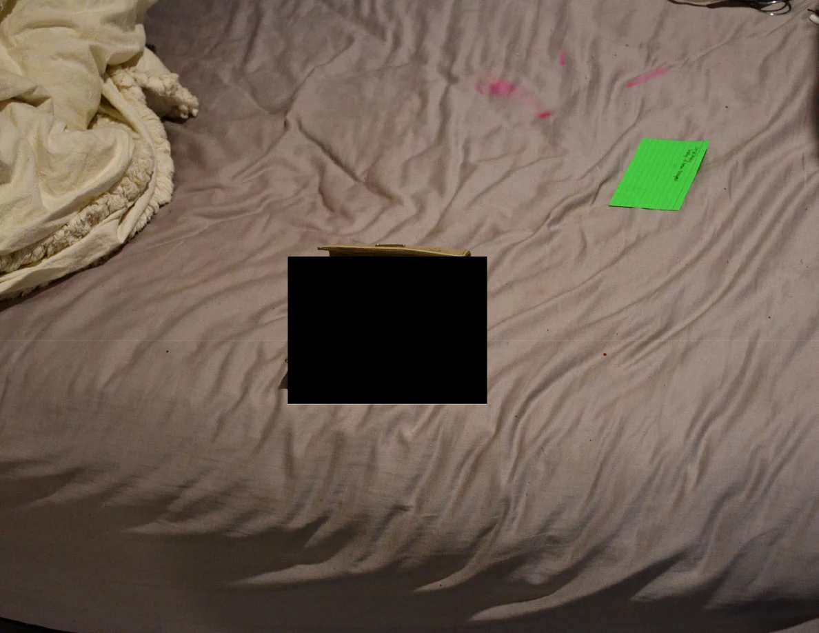 Bed surface with a partly redacted item, a green note and pink marks on the sheet.